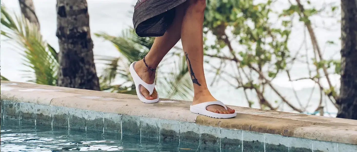 Femme marchant sur une bordure de piscine avec des sandales orthopédiques blanches Kidmi, entourée de palmiers.