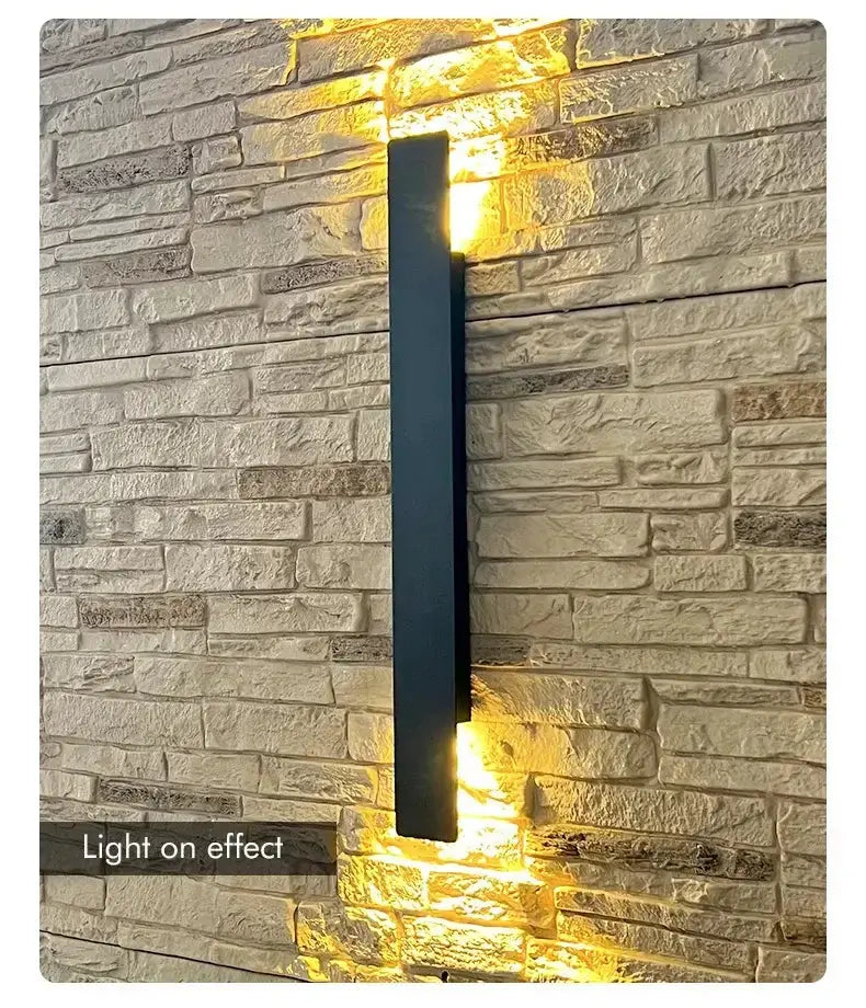 Applique murale LED noire allumée sur mur en pierre, créant un effet lumineux chaleureux.